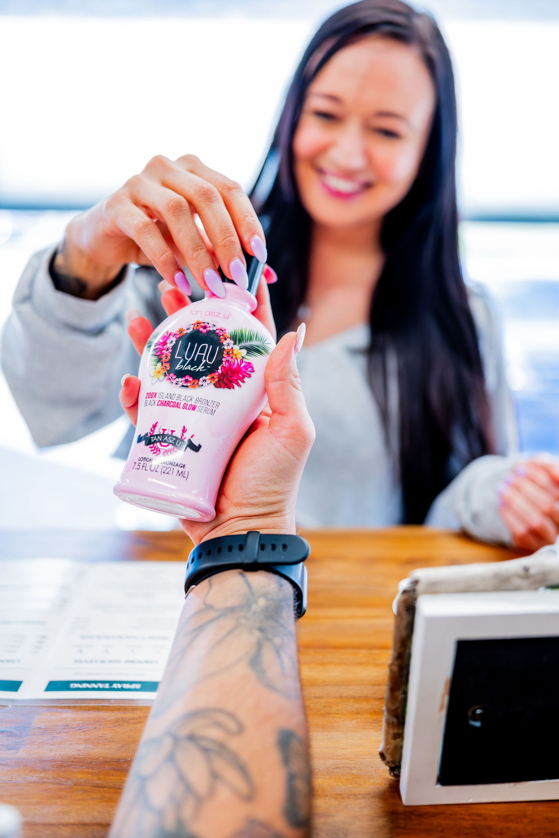 A woman with long dark hair smiles, reaching for a pink bottle from a tattooed arm. The setting is indoors.