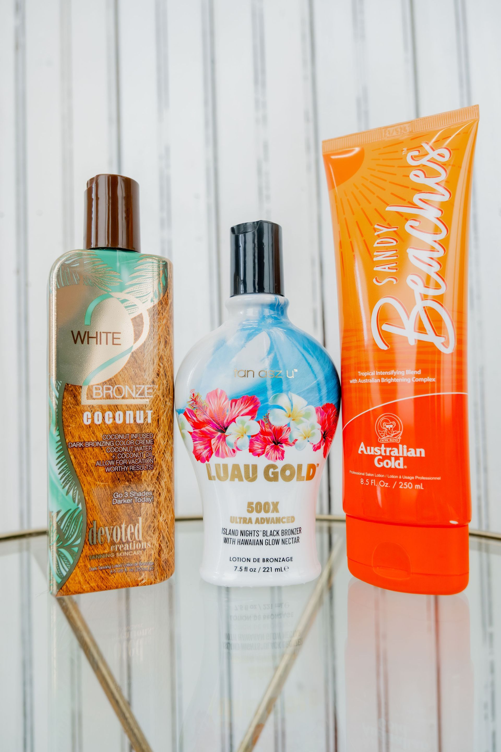 Three bottles of tanning lotion with brown, black, and orange caps stand on a reflective surface. The lotions feature tropical branding.