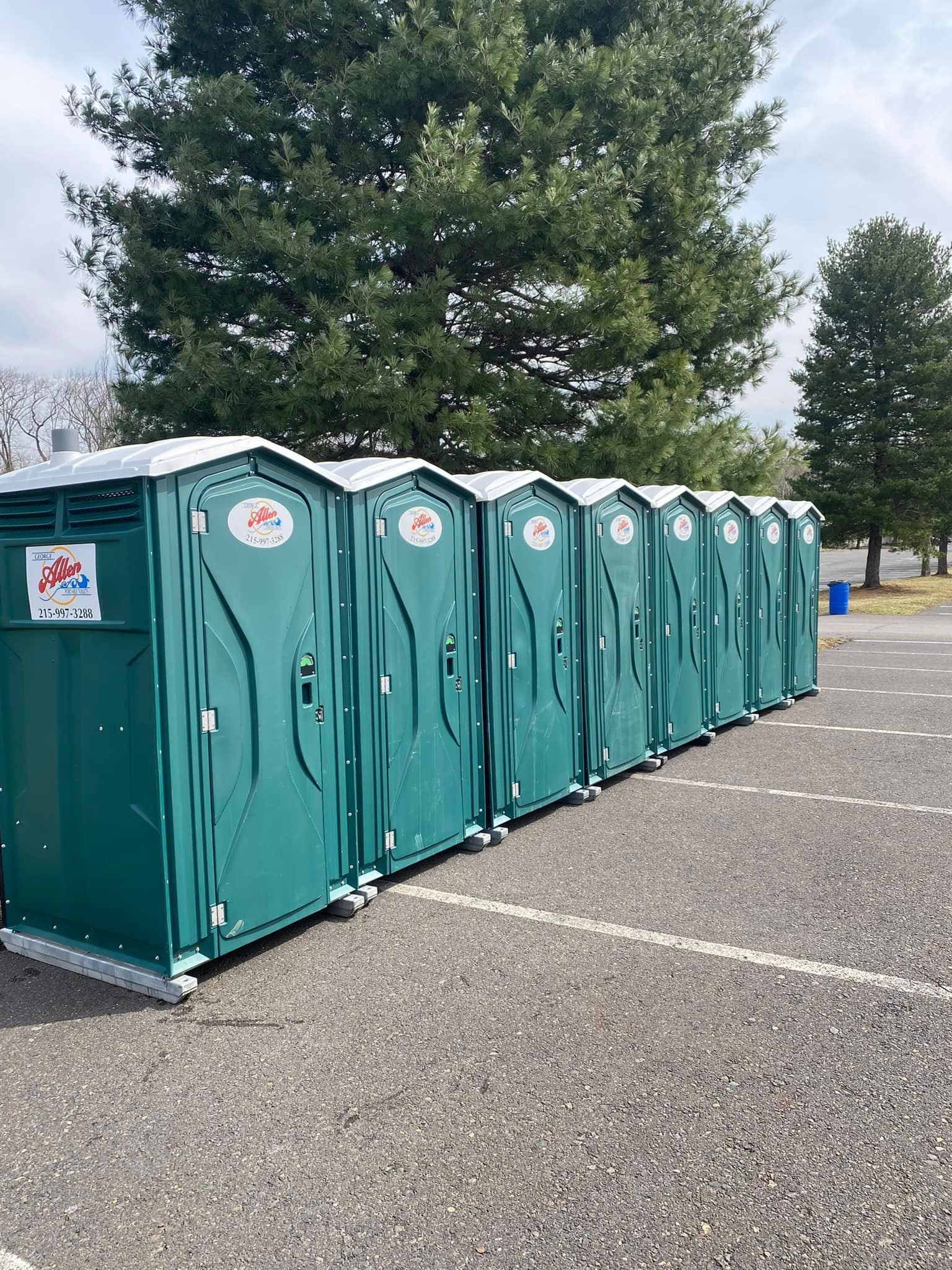 A row of eight green portable toilets lined up in an outdoor parking lot. A row of eight green portable toilets lined up in an outdoor parking lot.