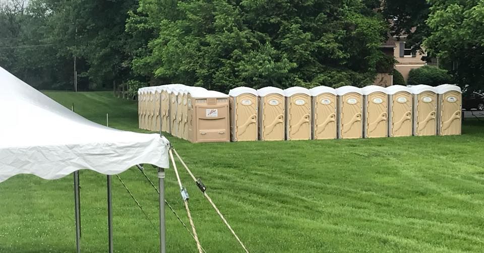 Row of tan portable toilets on a grassy field near a white tent and trees