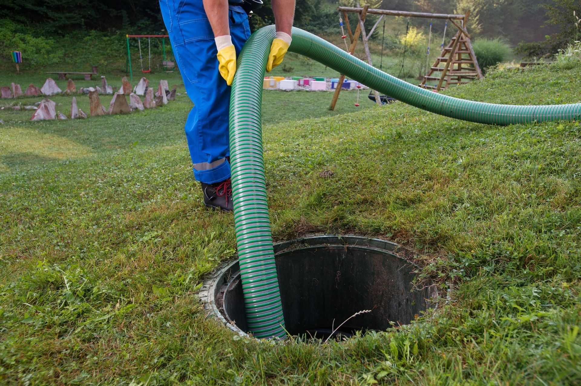 A person holding a large green hose inserted into an open septic tank.