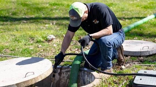 Worker wearing gloves and a cap, using equipment to clean or inspect an open manhole in a grassy area. Worker wearing gloves and a cap, using equipment to clean or inspect an open manhole in a grassy area.