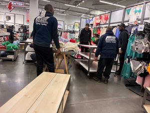 Four people moving store fixtures in a clothing store, with shelves and clothing racks visible.