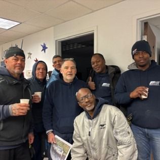 Group of men, some holding cups, inside a building. They are wearing various jackets and hats, smiling.