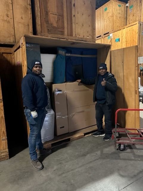 Two movers stand in a storage unit with packed boxes. One gives a thumbs-up.