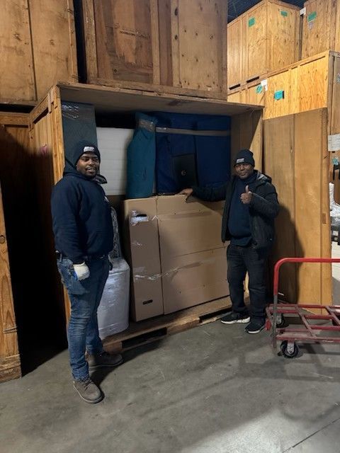 Two movers pose with packed boxes in a storage unit; one gives a thumbs up.