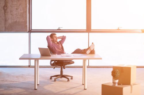 Person reclining in an office chair at a desk in a sunlit office, typing on a laptop.