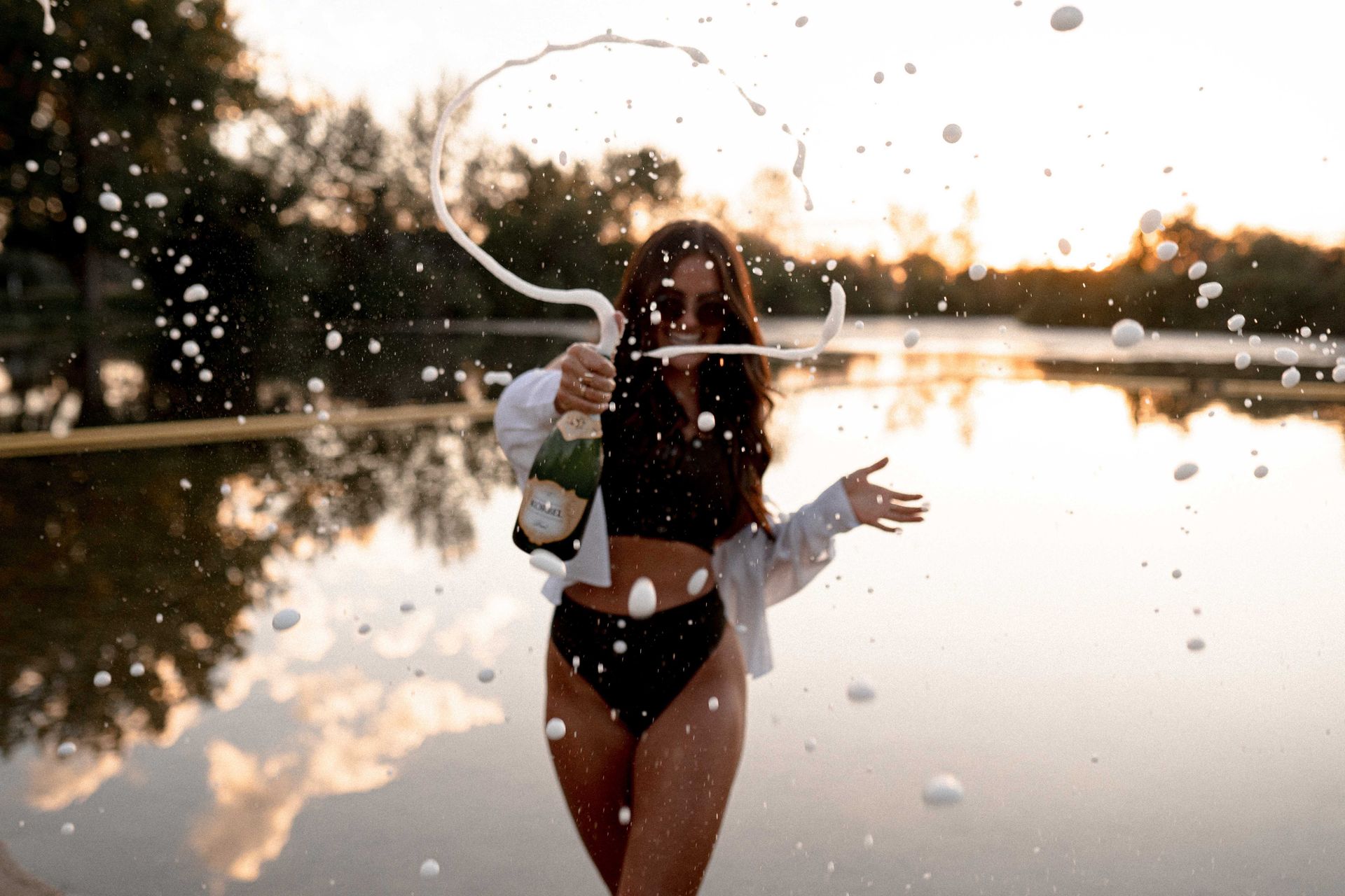 A woman in a bikini is holding a bottle of champagne and splashing it into the water.