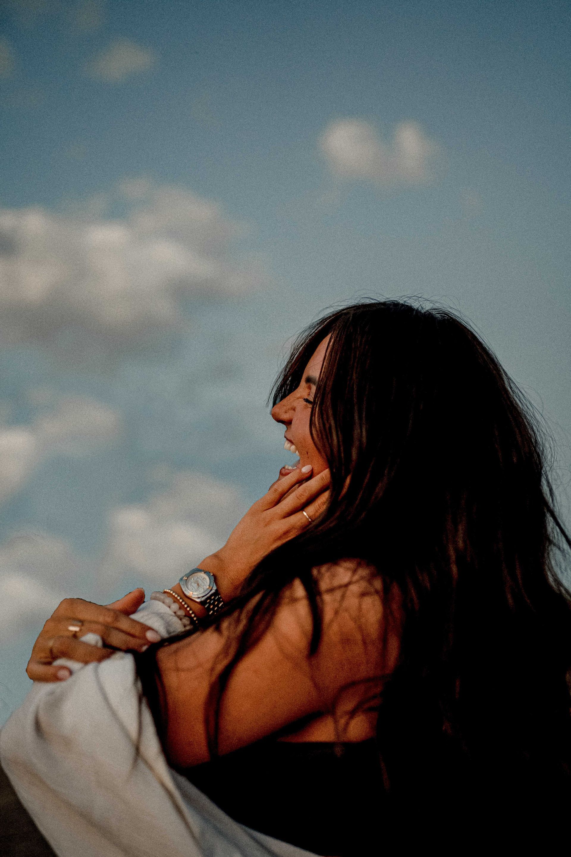 A woman with long hair is sitting in front of a blue sky with clouds.
