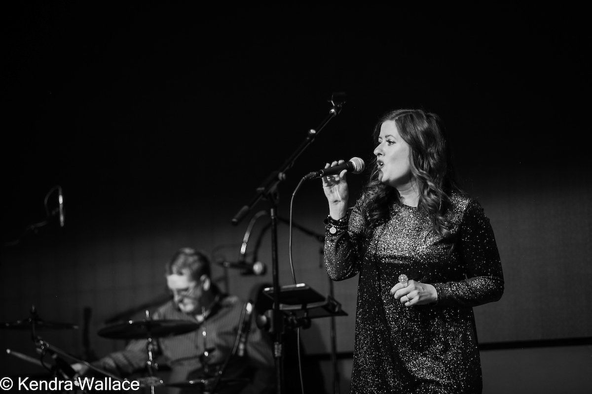 Woman singing into a microphone onstage, man playing keyboard in background, black and white photo.