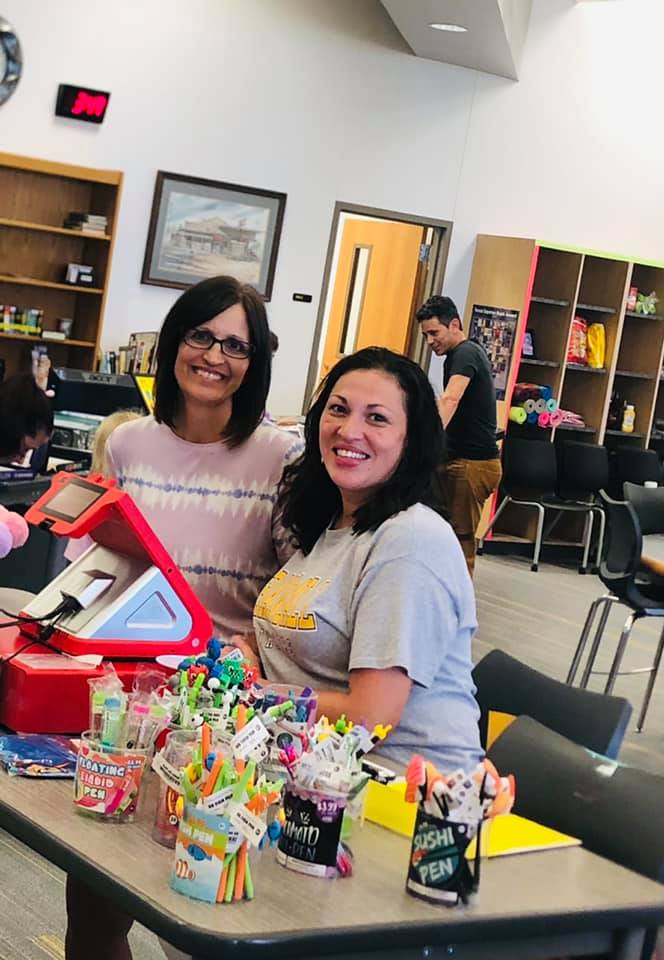 Two women smiling at a table with craft supplies and a cash register in a classroom.