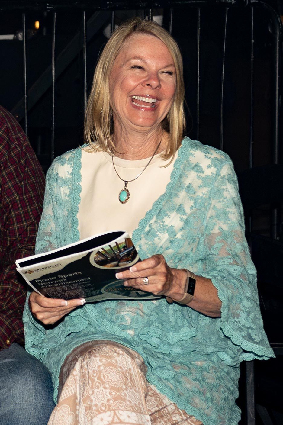 Woman laughing, holding a brochure, wearing a turquoise lace shawl and a turquoise pendant necklace.