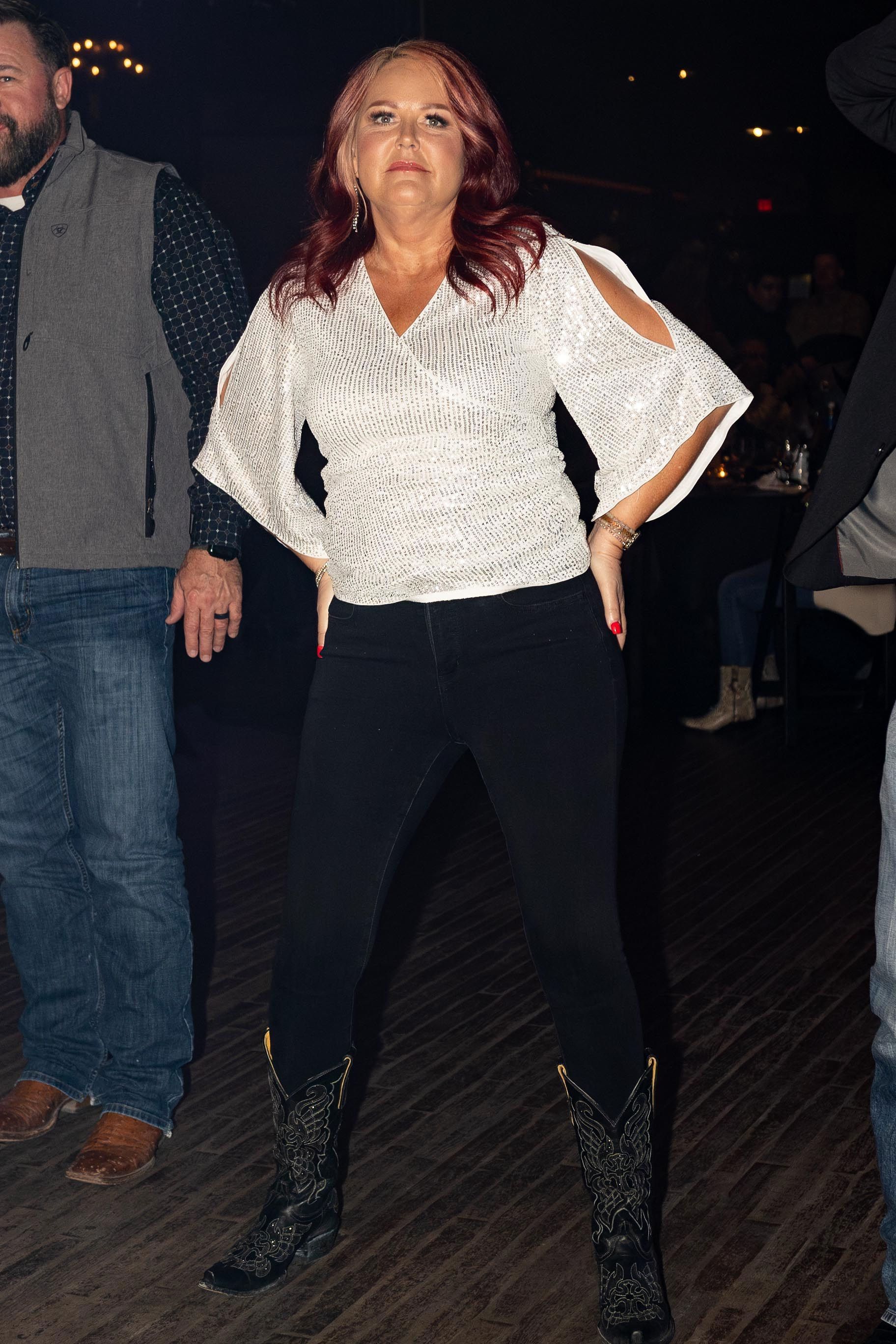 Woman with red hair poses in a white top, black jeans, and boots, hands on hips, at a venue.