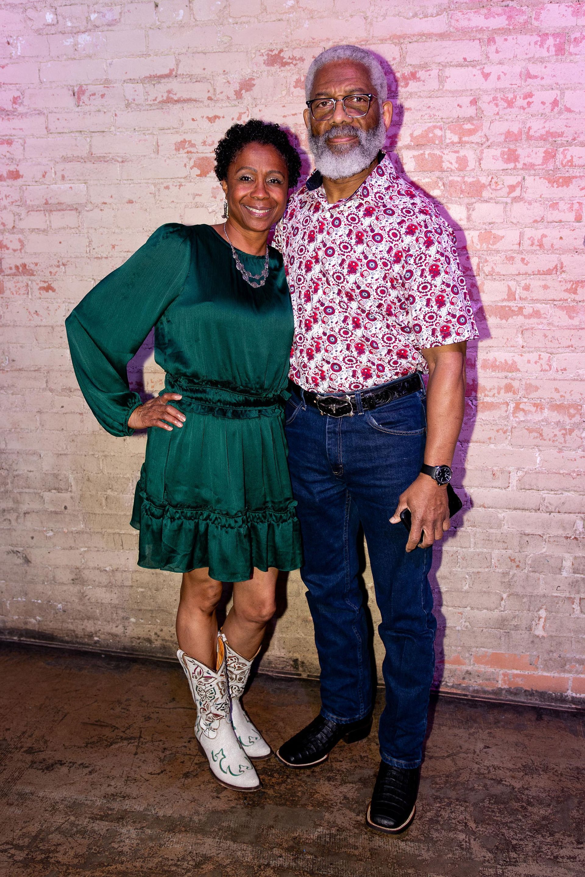 Couple posing near a brick wall. Woman in green dress and cowboy boots; man in floral shirt and jeans.