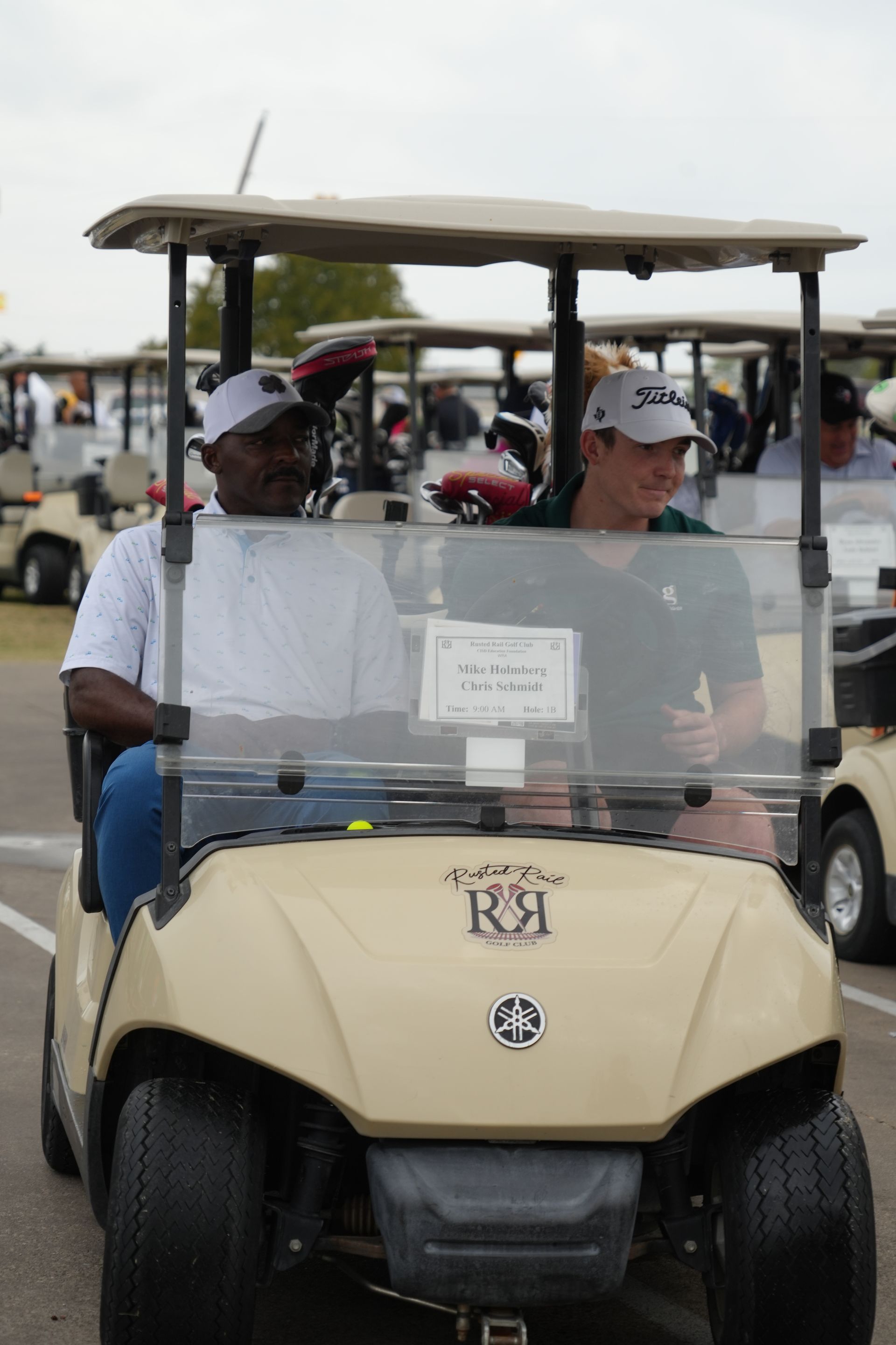 Two men in a golf cart at an outdoor event. One is Black, the other is white. Many other golf carts are present.