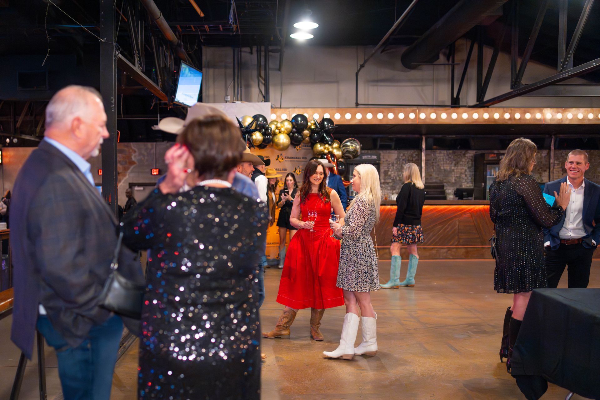 People at an event in a large room. Woman in red dress, woman in sequin top, balloons, bar, and tables.