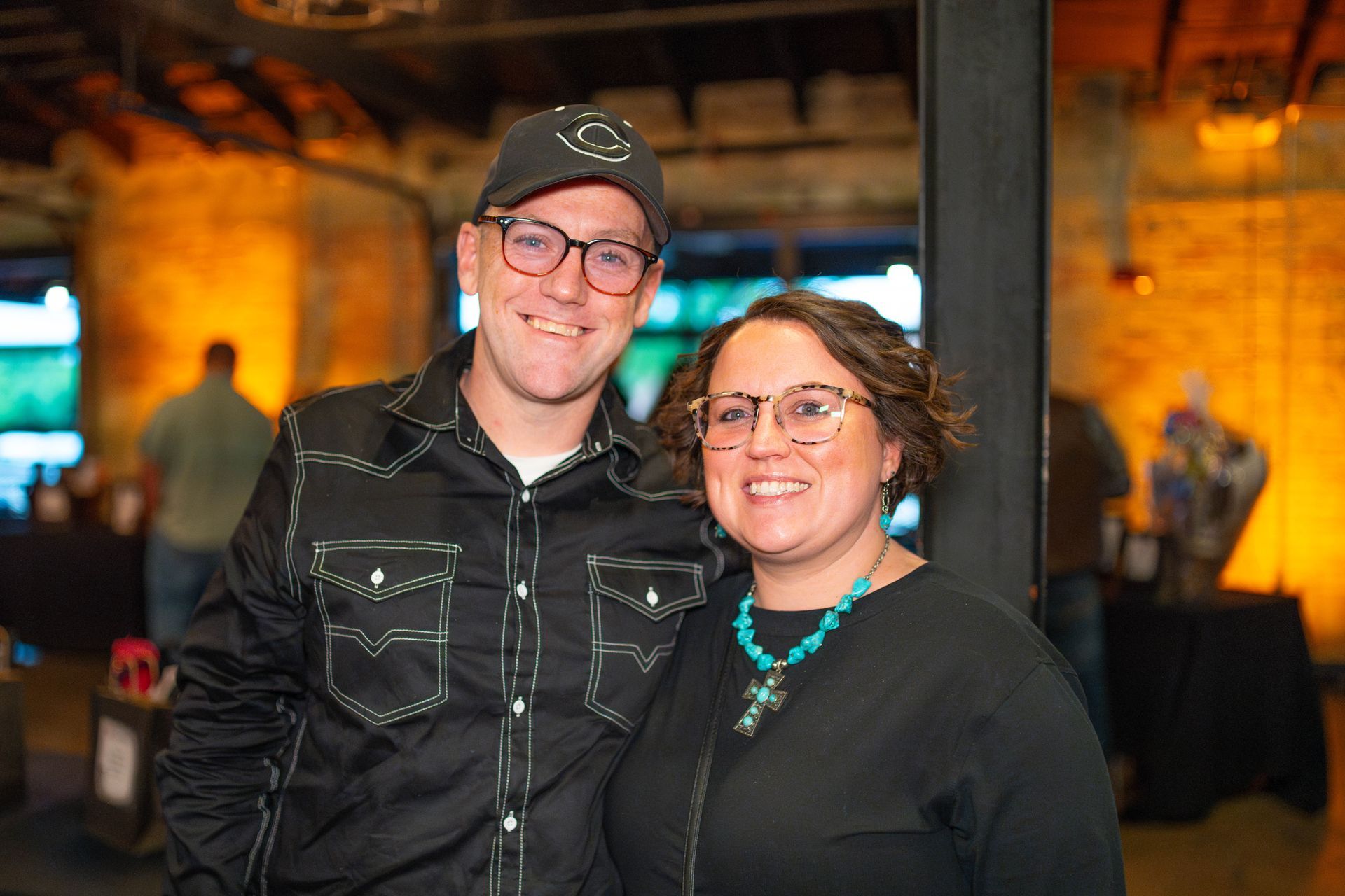 Man and woman smiling, posing together inside a building. Man in a black shirt and hat. Woman wears glasses and a turquoise necklace.