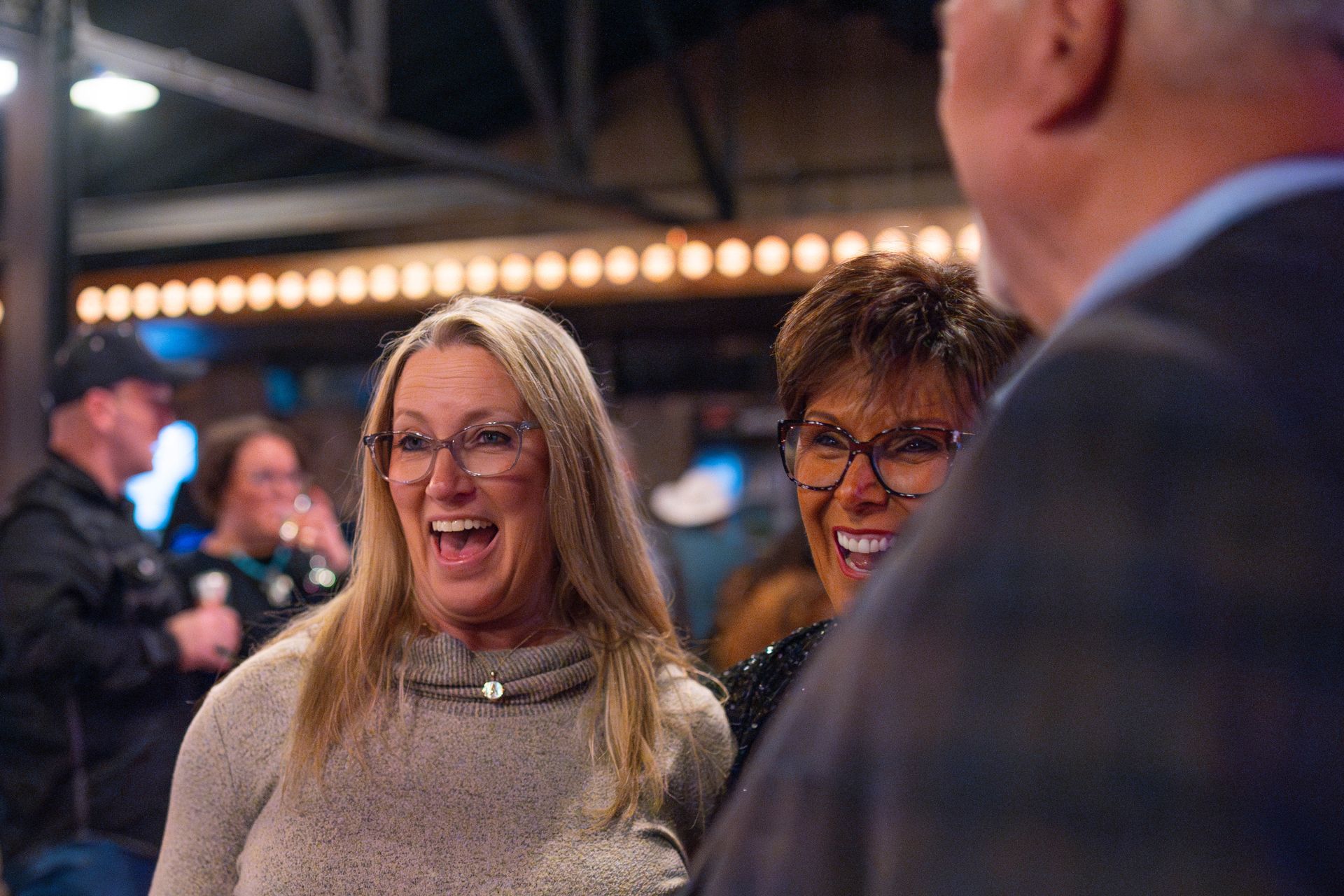 Two women with glasses laughing; lit-up background, man in foreground.