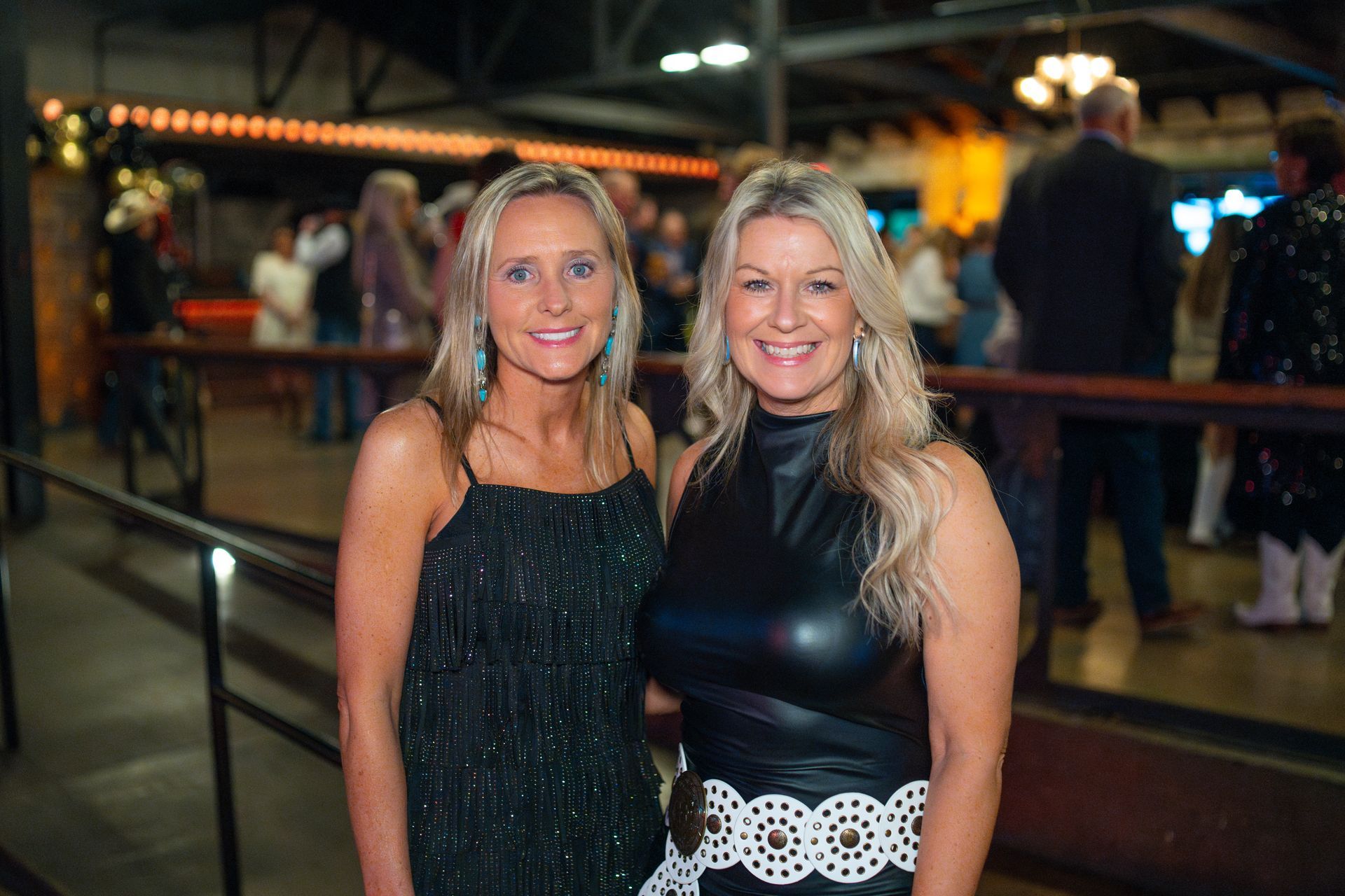 Two women smiling, posing indoors with others blurred in background. One wears a black dress, the other a leather-look top.