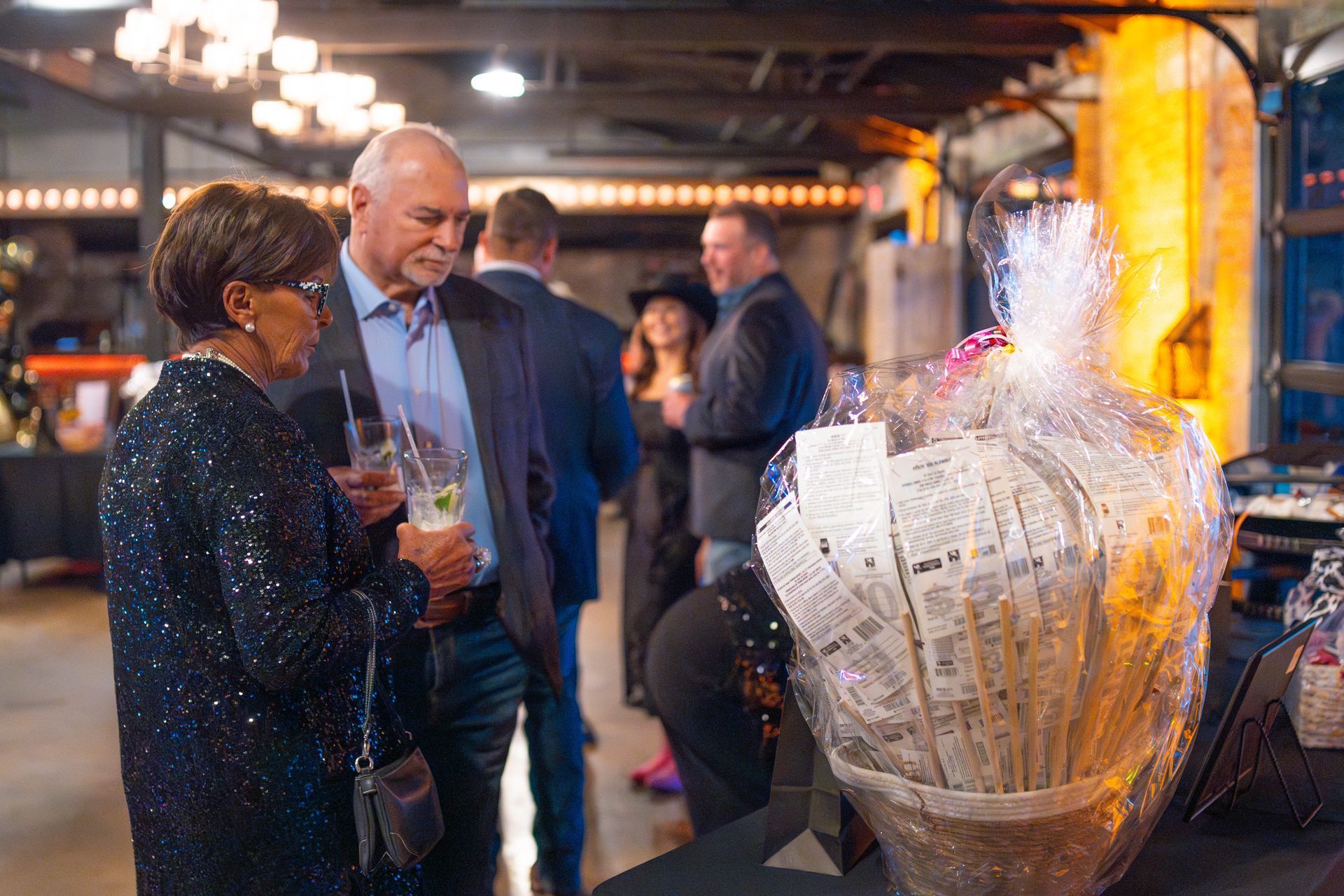 Couple at an event, looking at a raffle basket. Dark interior, other guests visible.
