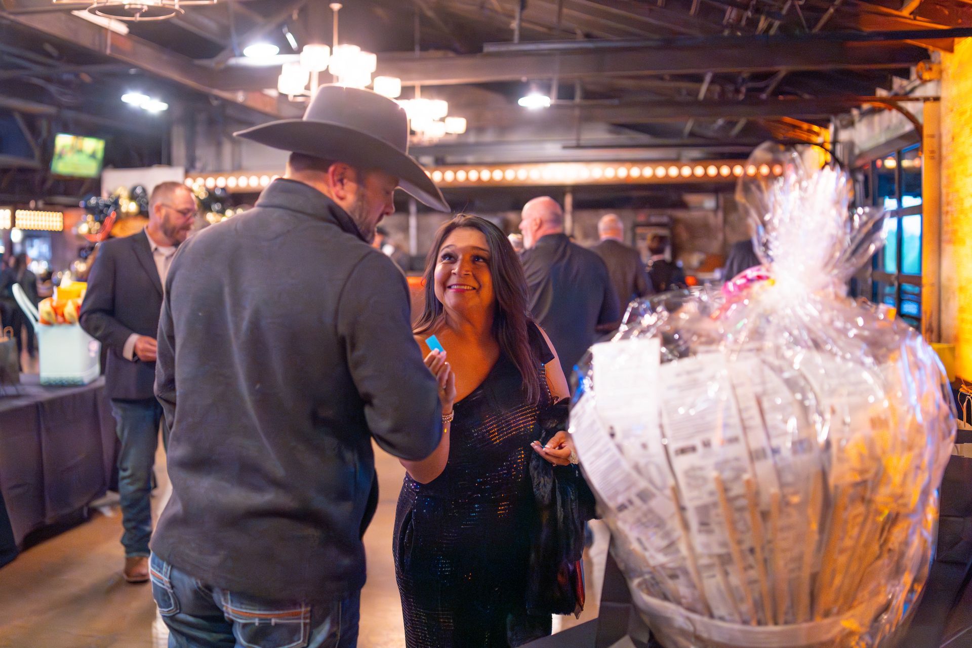 Man in cowboy hat and woman in sparkly dress talking, basket in the foreground at an event.