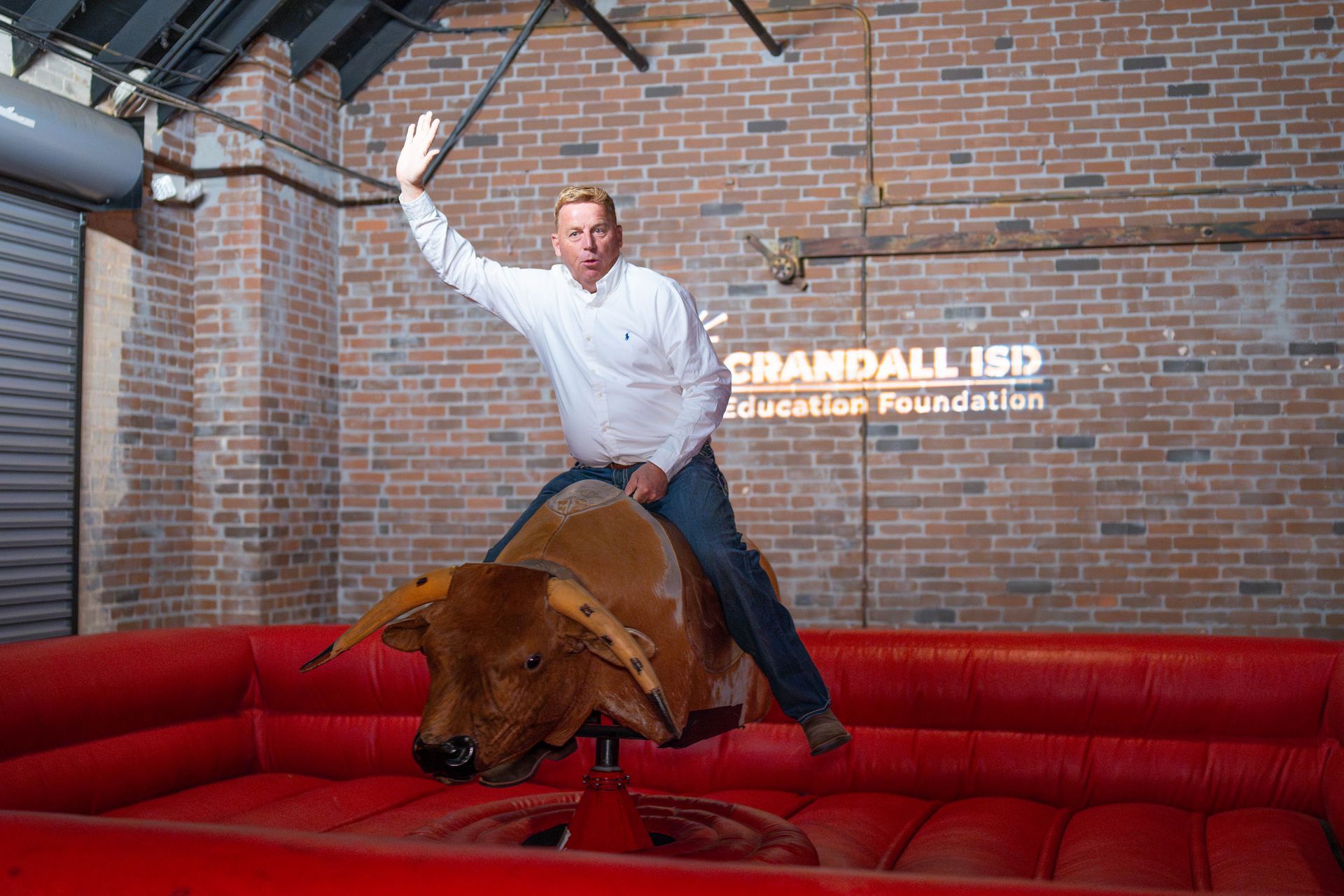 Man riding a mechanical bull, waving, in front of a brick wall with 