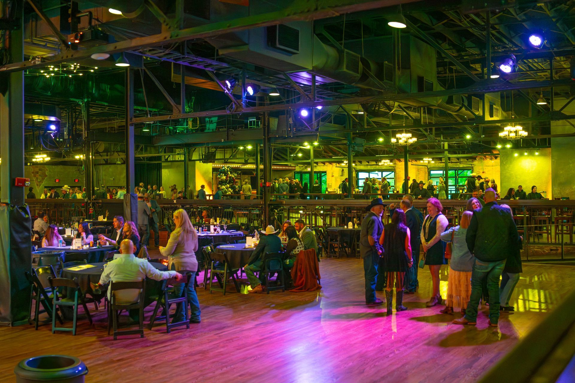 People seated and standing in a large, brightly lit dance hall with tables and a bar.