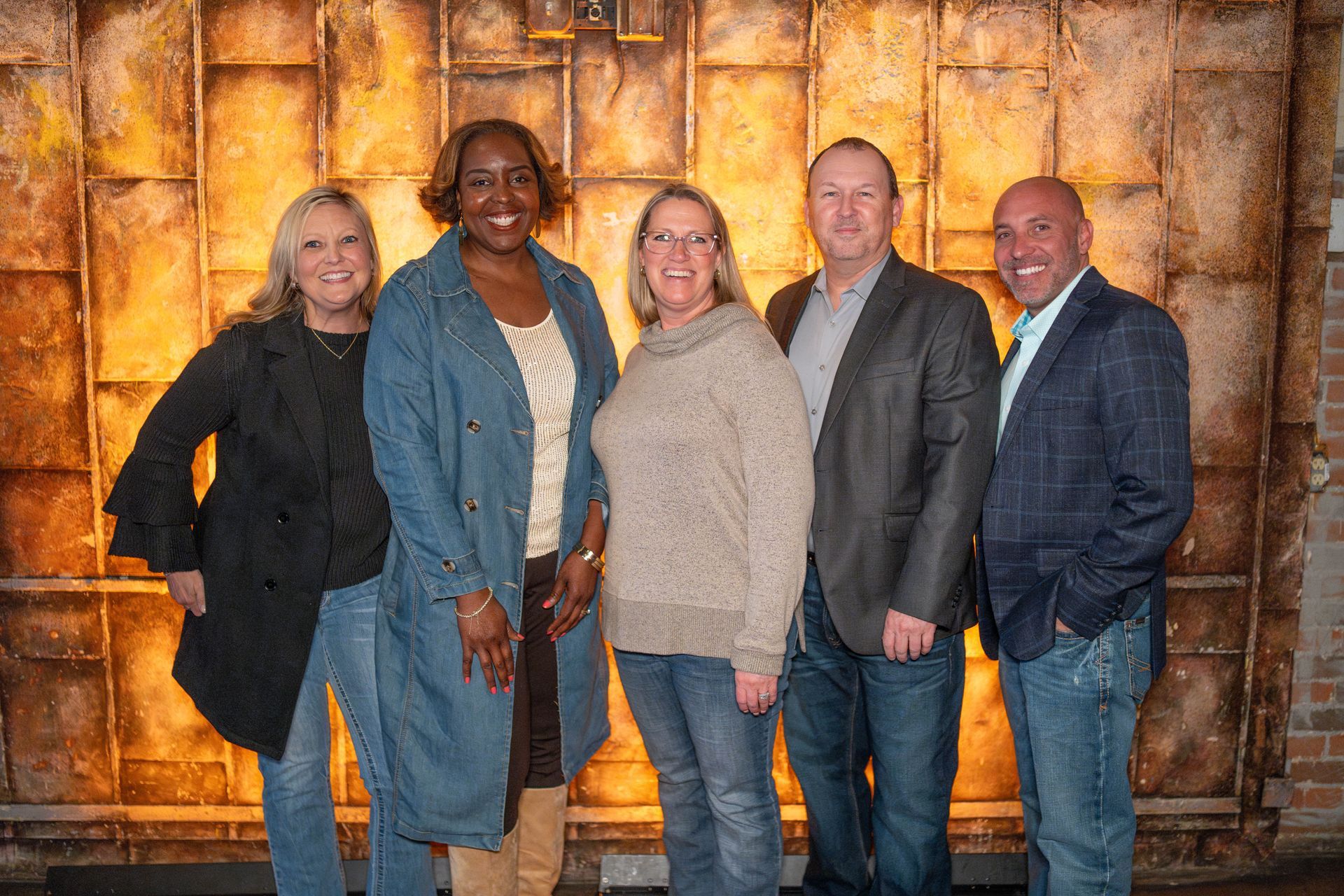 Five people pose in front of a textured gold wall. Two women on the left, three on the right, smiling.