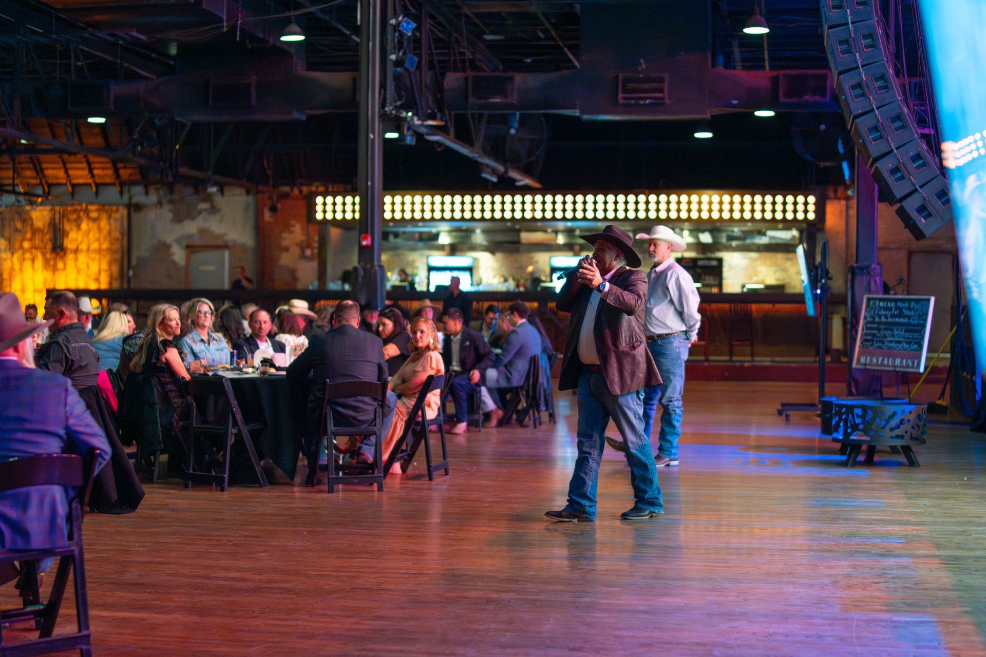 Cowboy hat-wearing men speaking to a seated audience at a dimly lit event in a venue.