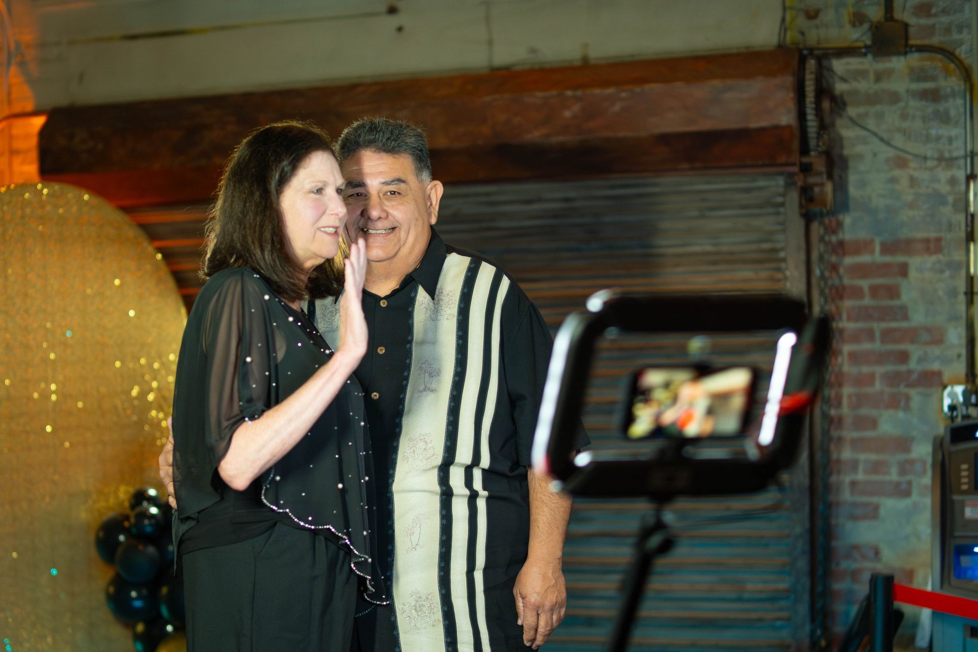 Couple posing for photo booth, woman waving, man smiling; industrial backdrop.