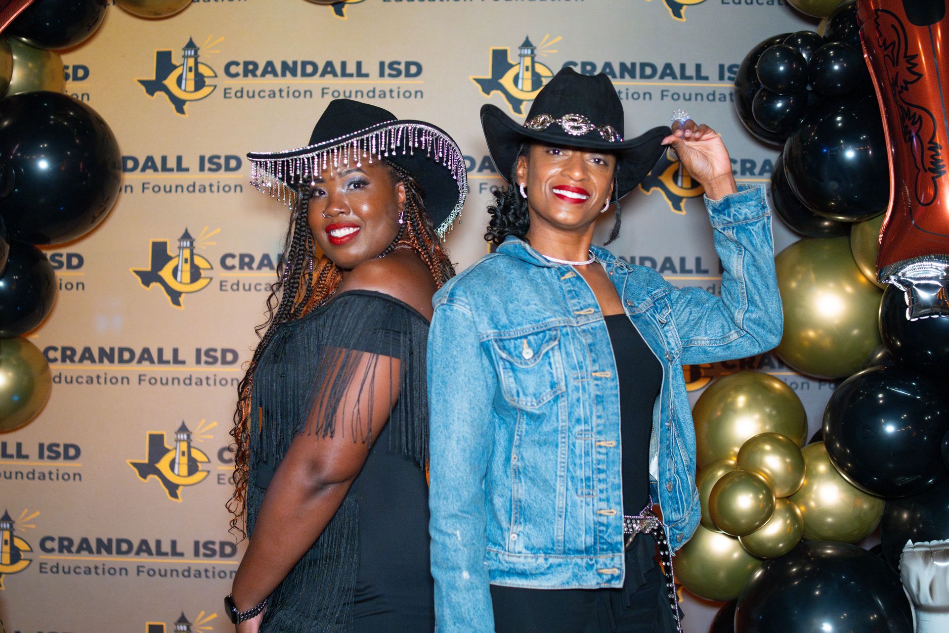 Two women in cowboy hats pose, smiling, at an event with balloons and a Crandall ISD backdrop.
