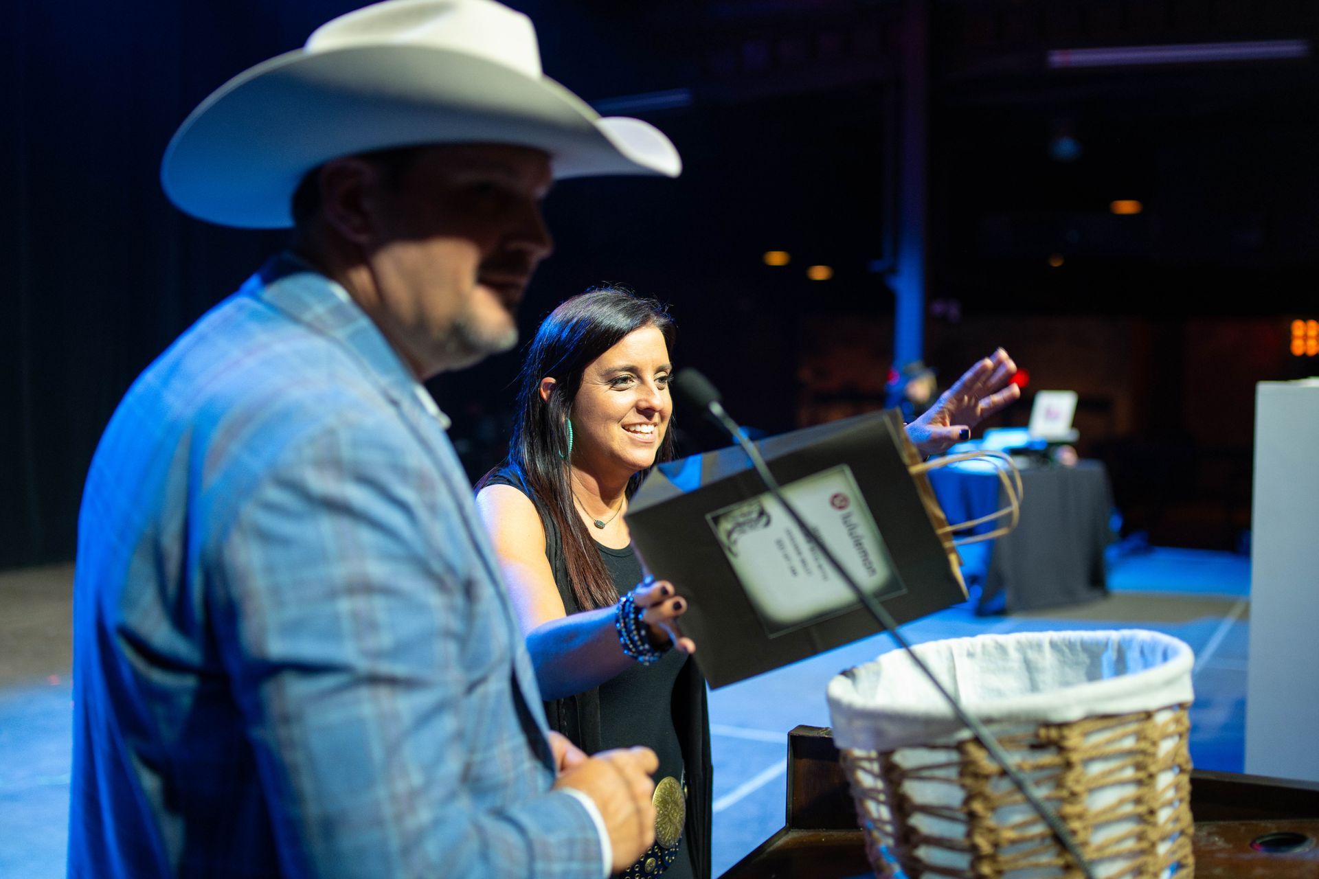 Man in cowboy hat and woman at a podium, holding a framed document and gesturing, near a wicker basket.