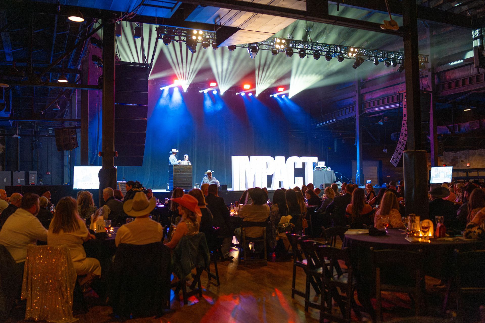 Audience seated at tables, stage with IMPACT sign and speakers, lights in a large hall.