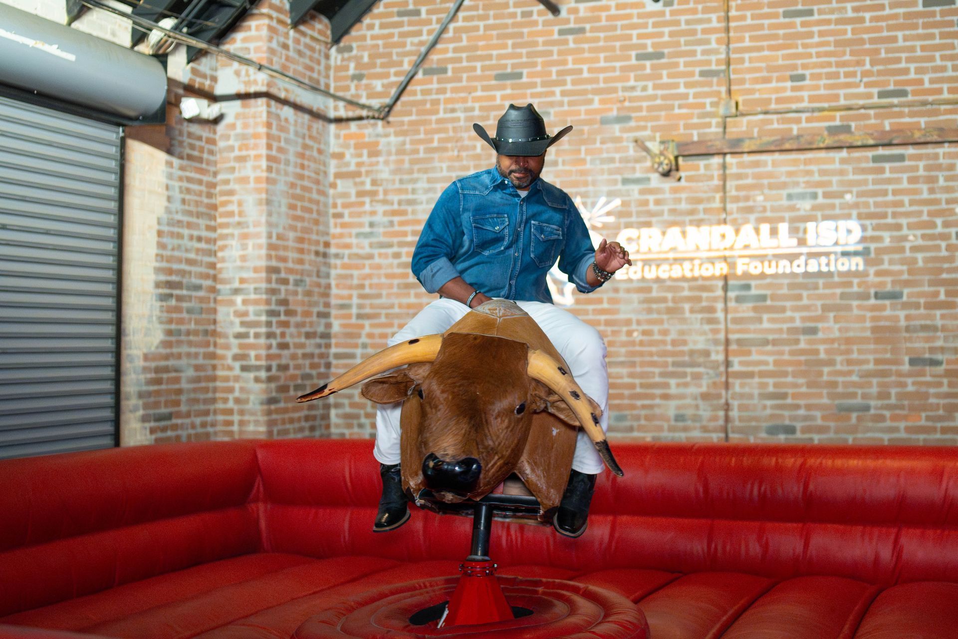 Man in cowboy hat riding a mechanical bull in a red-padded arena against a brick wall.
