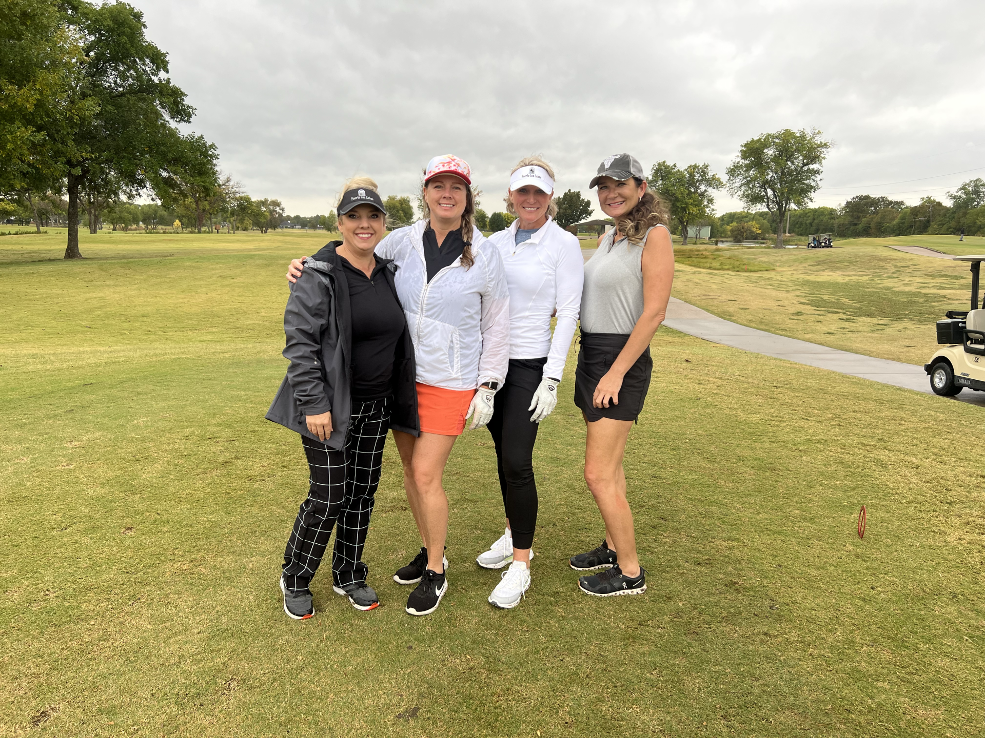 Four women pose on a golf course, smiling. They wear golf attire. Overcast day.
