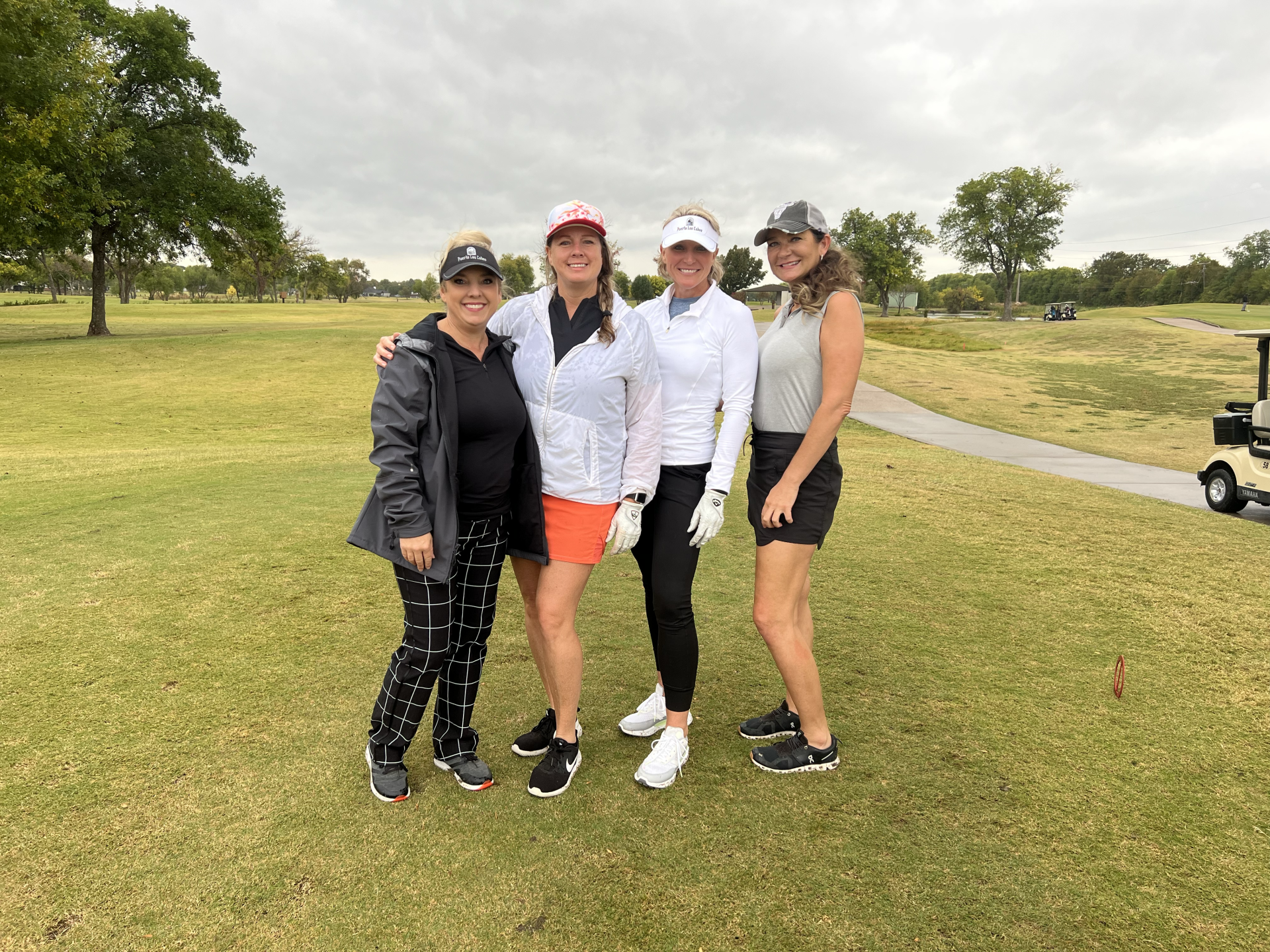Four women in golf attire pose on a green course under cloudy skies.