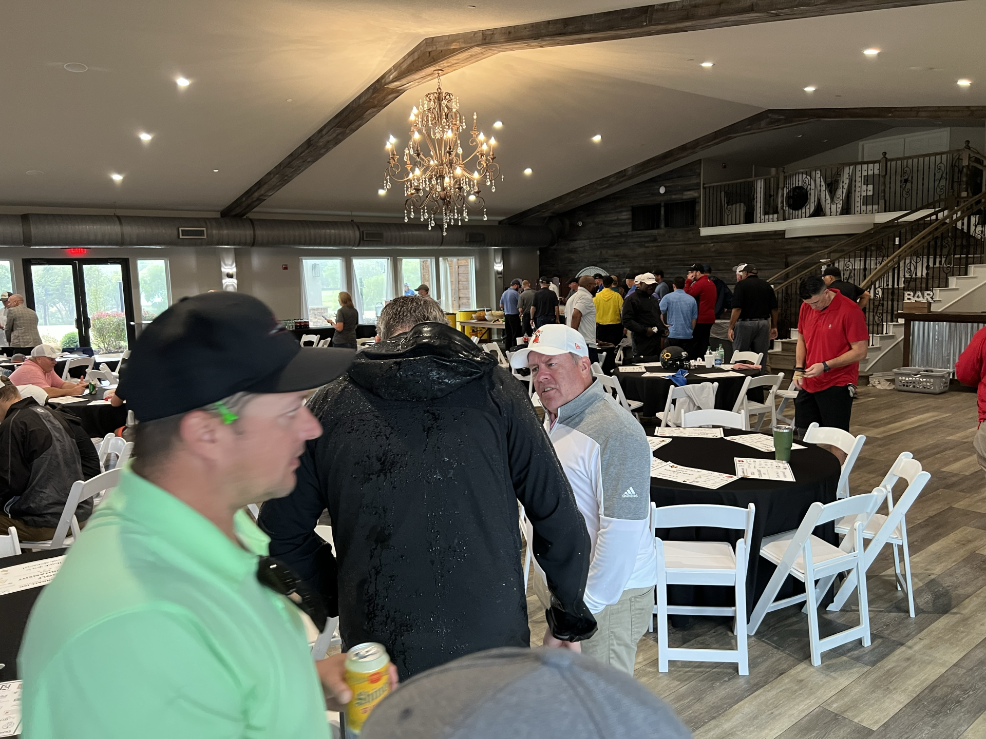 People in a large room with tables, a chandelier, and a staircase. Some wear golf attire.