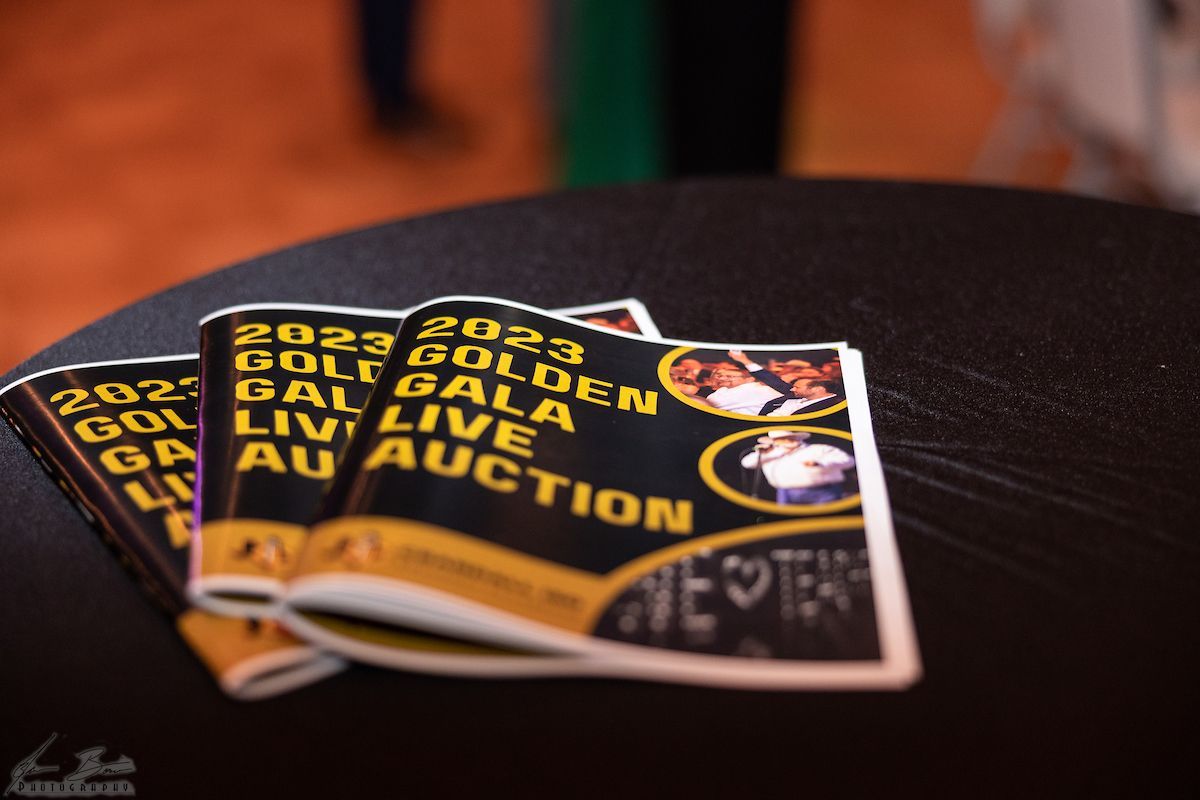 Several 2023 Golden Gala Live Auction booklets on a black table with gold and black cover design.