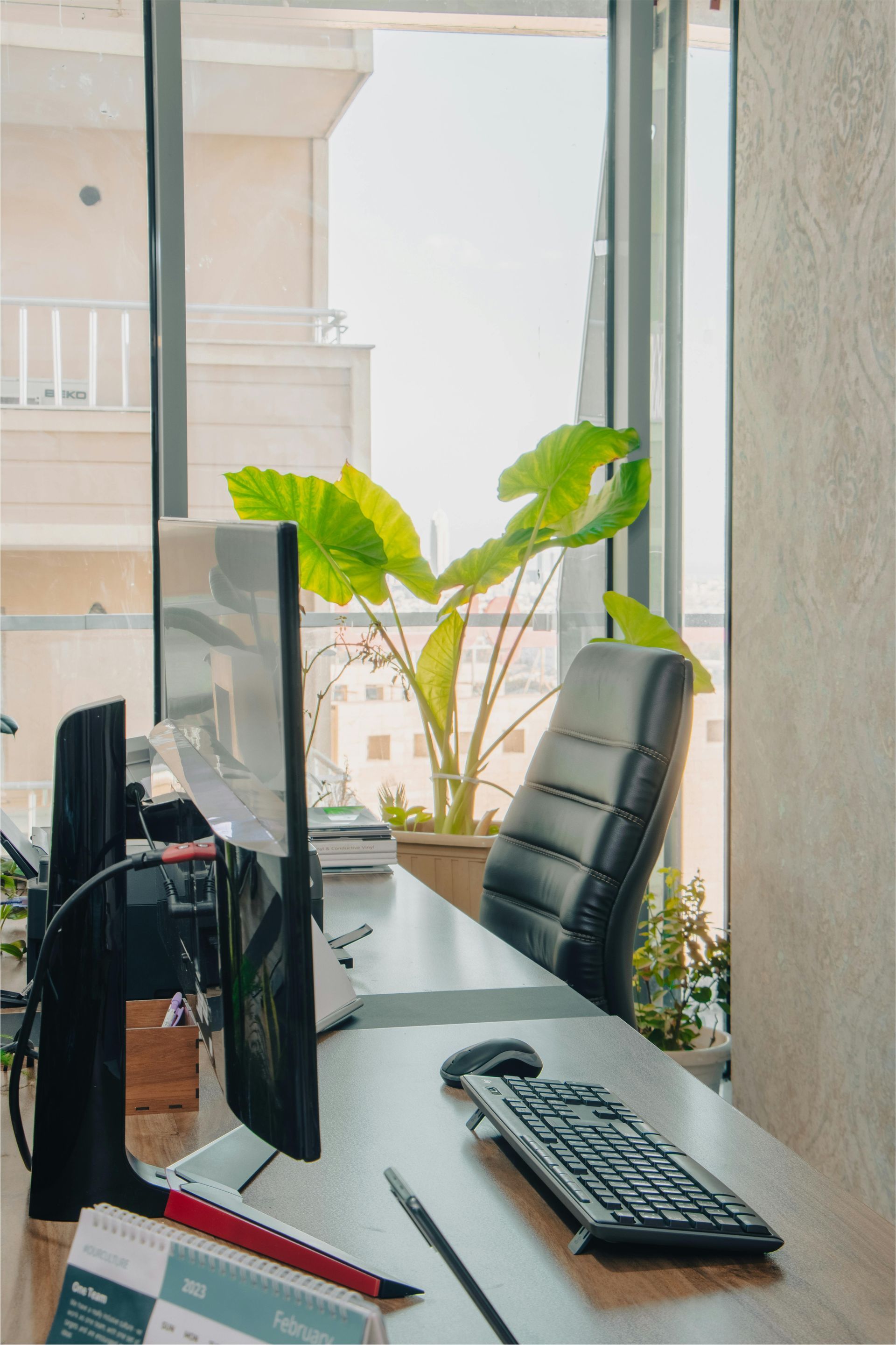 Office desk by a window with a computer monitor, keyboard, chair, and potted plant.