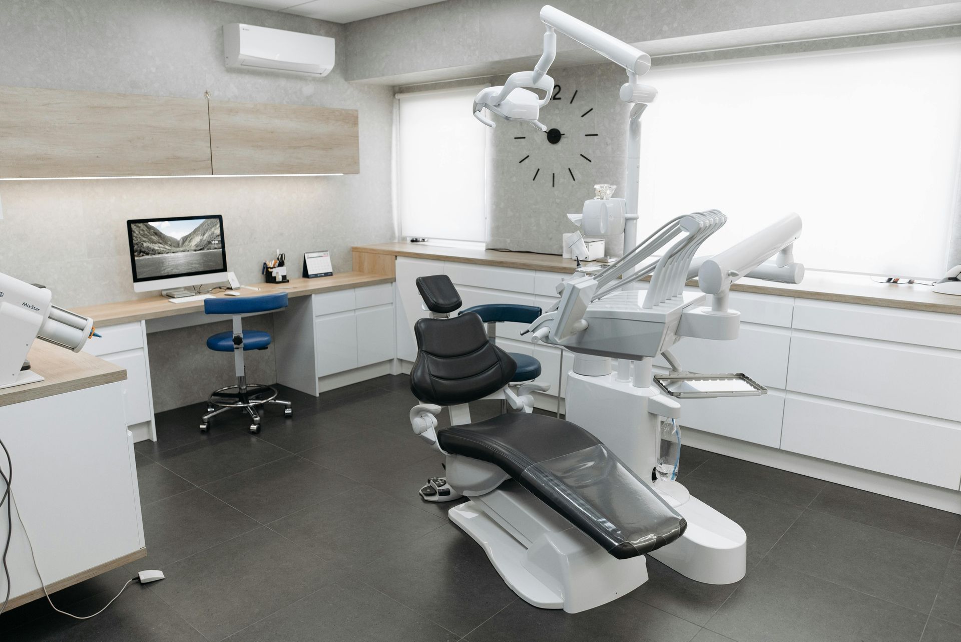 Modern dental exam room with a reclining chair, overhead light, computer desk, and white cabinets.