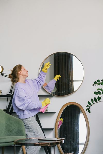 Person dusting a round mirror in a bright living room with purple gloves and a lavender sweater