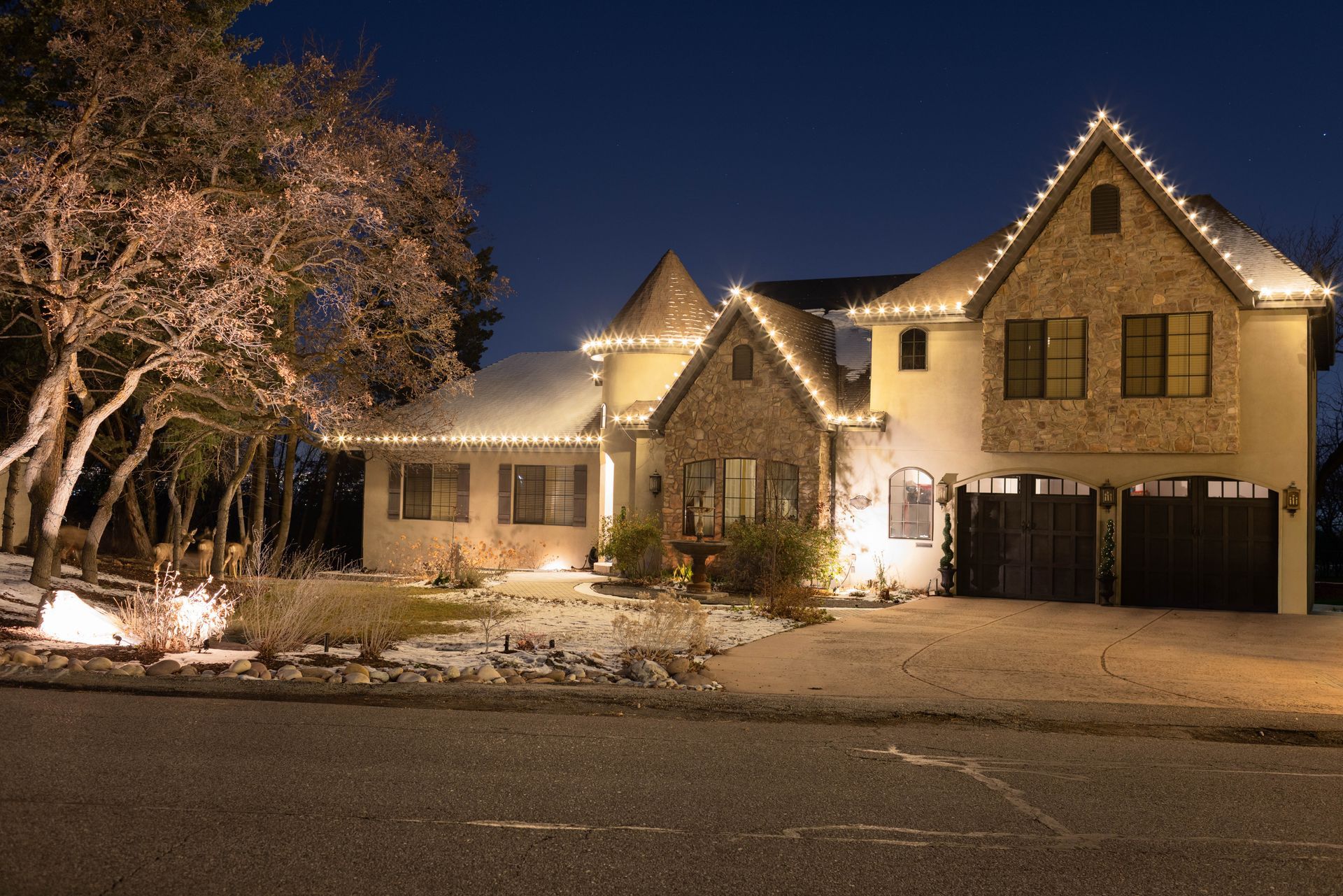 Snow-covered house decorated with warm white lights at night, with a lit tree in the front yard.