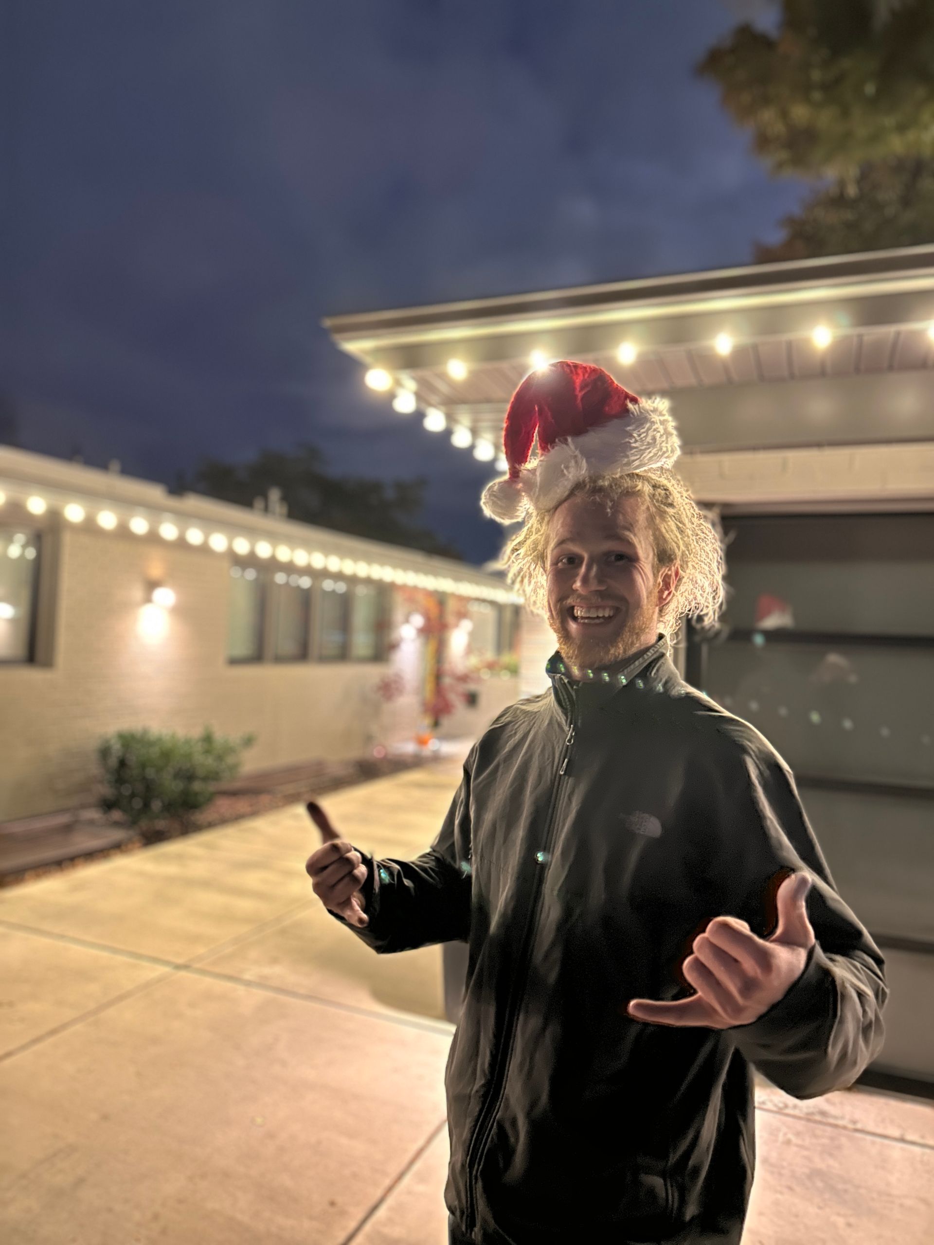 Person in a Santa hat and black jacket stands smiling in a lit driveway at night.