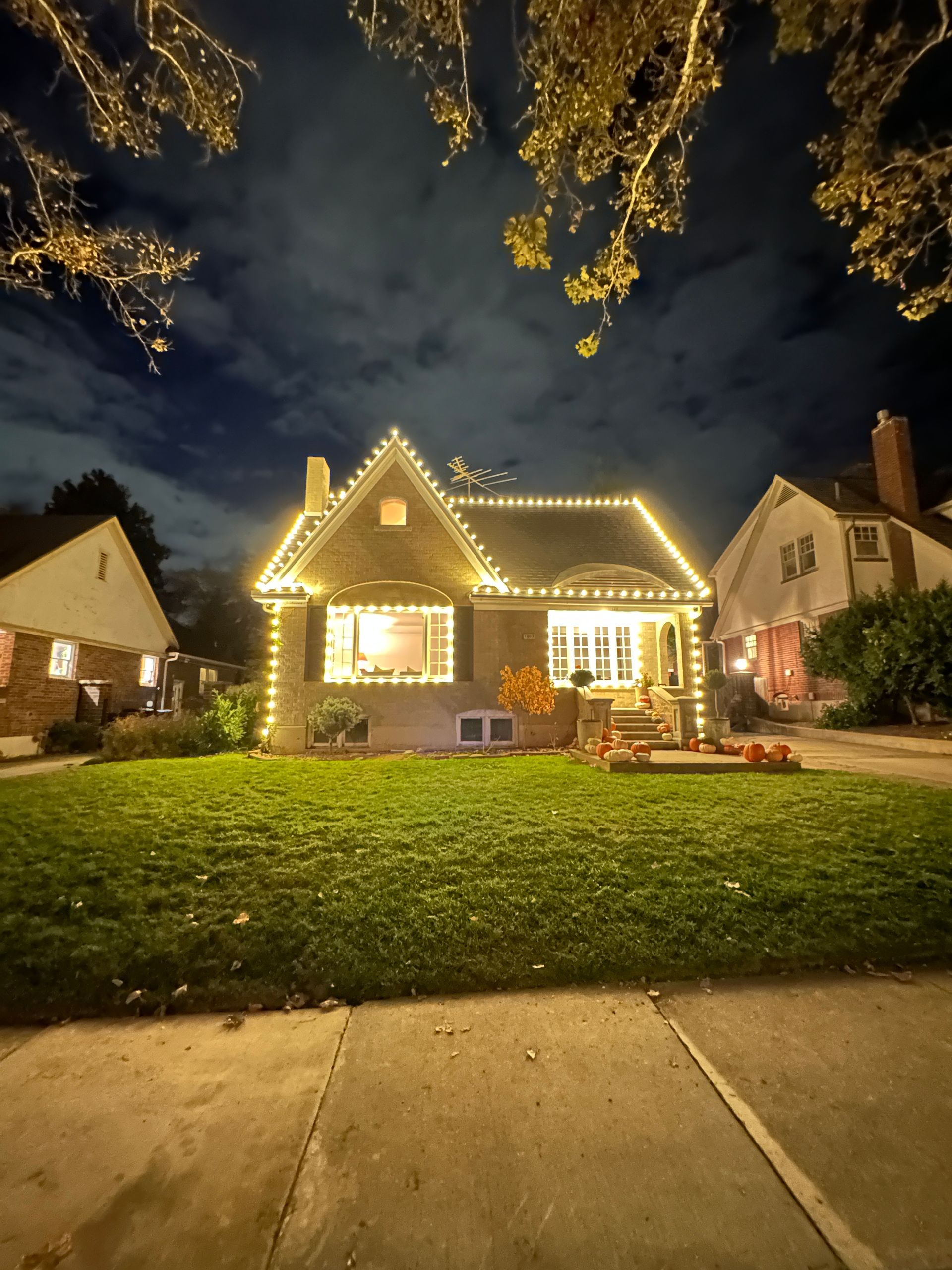 House with string lights on the roof, lawn in front, and two neighboring houses. Night scene.