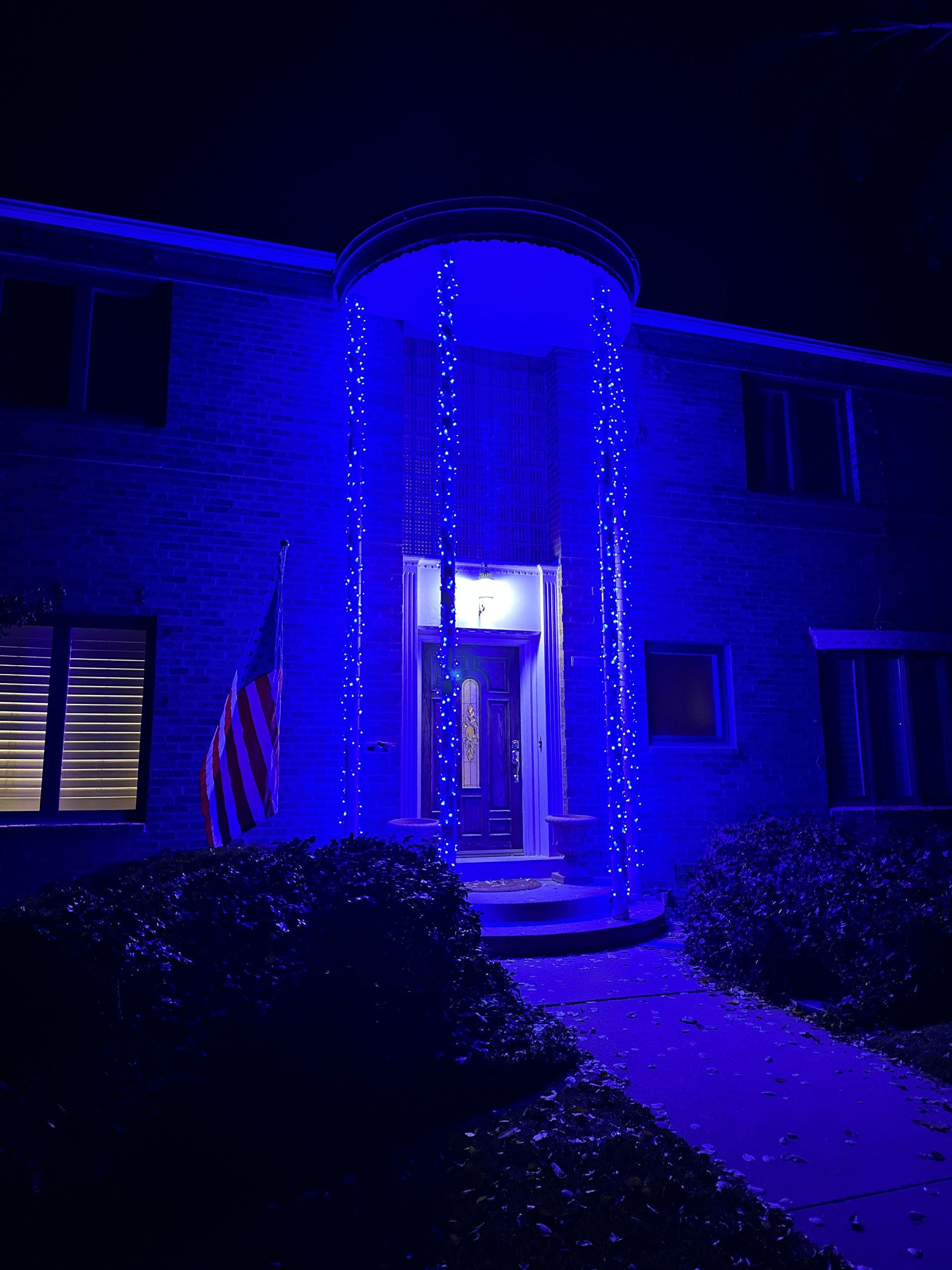 A two-story house with blue lights draped over the entrance, illuminated at night. An American flag is visible.