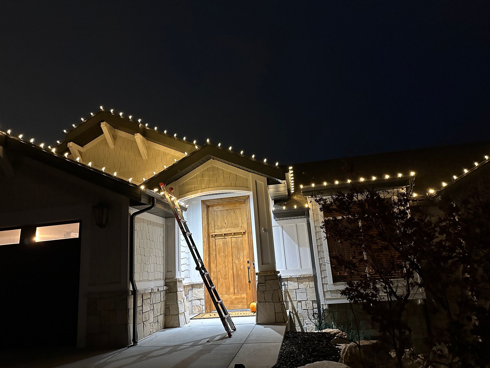 House at night with Christmas lights on the roof. Ladder leans against the front door, snow on the ground.