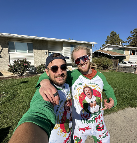 Two men in matching Elf-themed pajamas smile outdoors. One has sunglasses and a hat; the other, sunglasses and a pointing gesture.