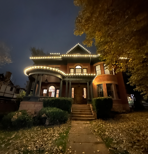 A large, illuminated house at night, with lights outlining the roof and porch.