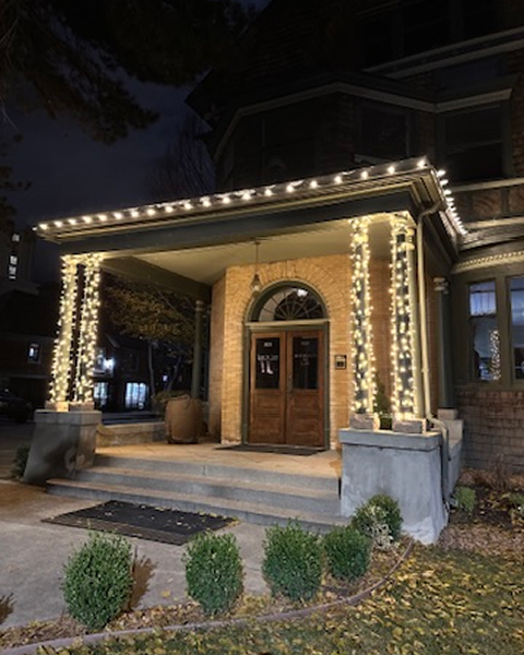Lit-up porch of a brick building at night. String lights adorn the columns and roof, with bushes and a door.