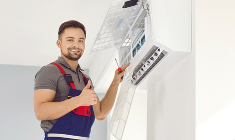 Technician in gray shirt and red overalls adjusting a wall-mounted electrical panel with a screwdriver